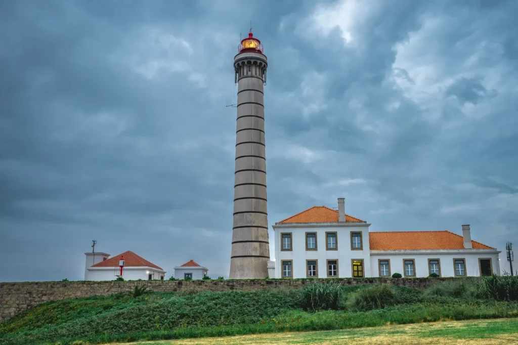 Farol branco debruçado sobre o mar agitado ao entardecer, com a lanterna acesa e névoa na base da torre