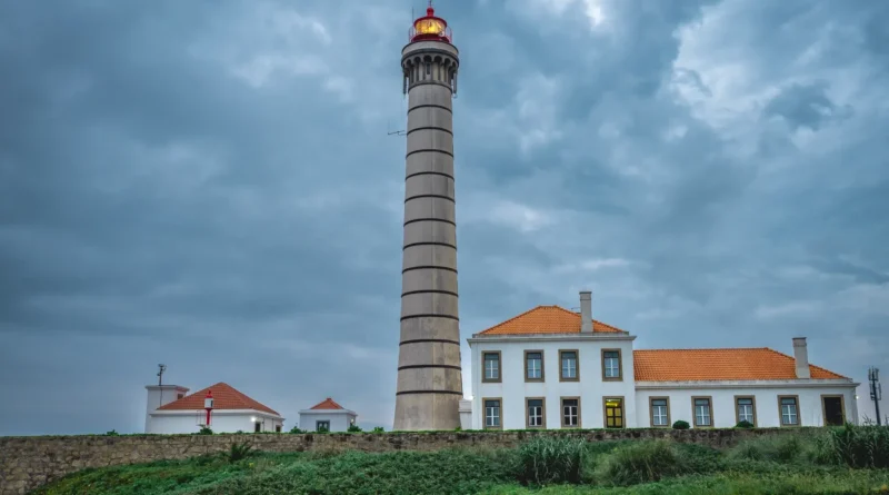 Farol branco debruçado sobre o mar agitado ao entardecer, com a lanterna acesa e névoa na base da torre