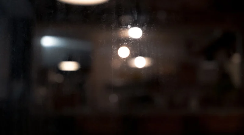 Recepção de academia vazia à noite, com luz amarelada baixa, balcão de madeira e chuva escorrendo na janela, ambiente silencioso frequentado apenas por adultos