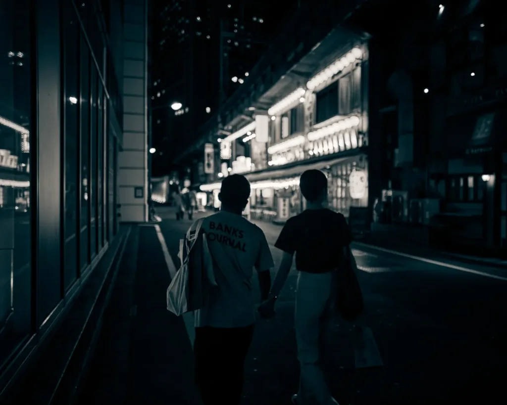 Dois adultos caminham juntos por uma rua iluminada à noite, cada um com uma mochila de ginástica, sorrisos discretos e clima de cumplicidade.