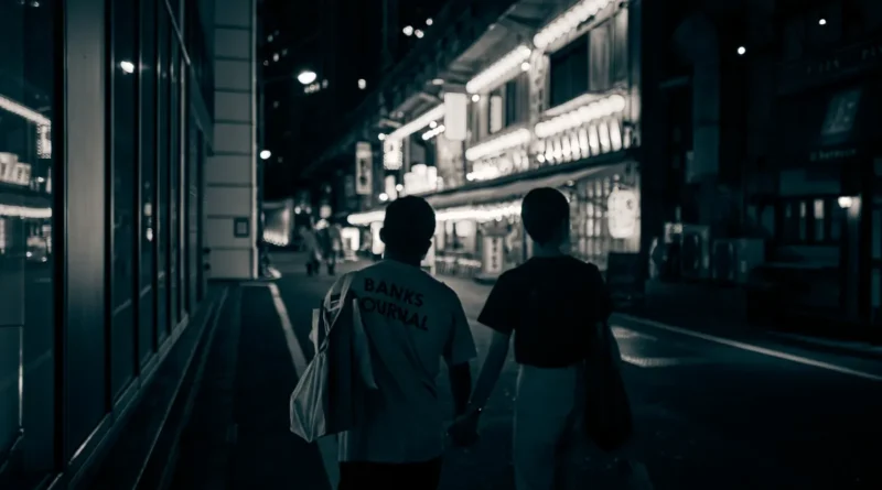 Dois adultos caminham juntos por uma rua iluminada à noite, cada um com uma mochila de ginástica, sorrisos discretos e clima de cumplicidade.