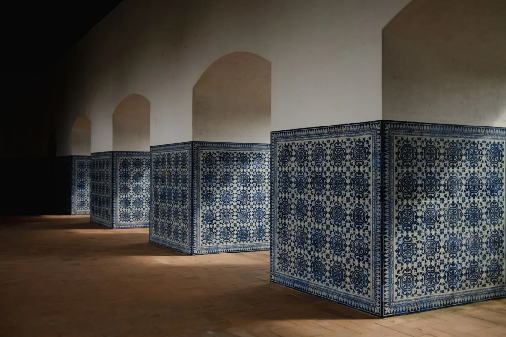 Duas pessoas adultas observam juntas um painel de azulejos restaurado numa oficina iluminada em Lisboa